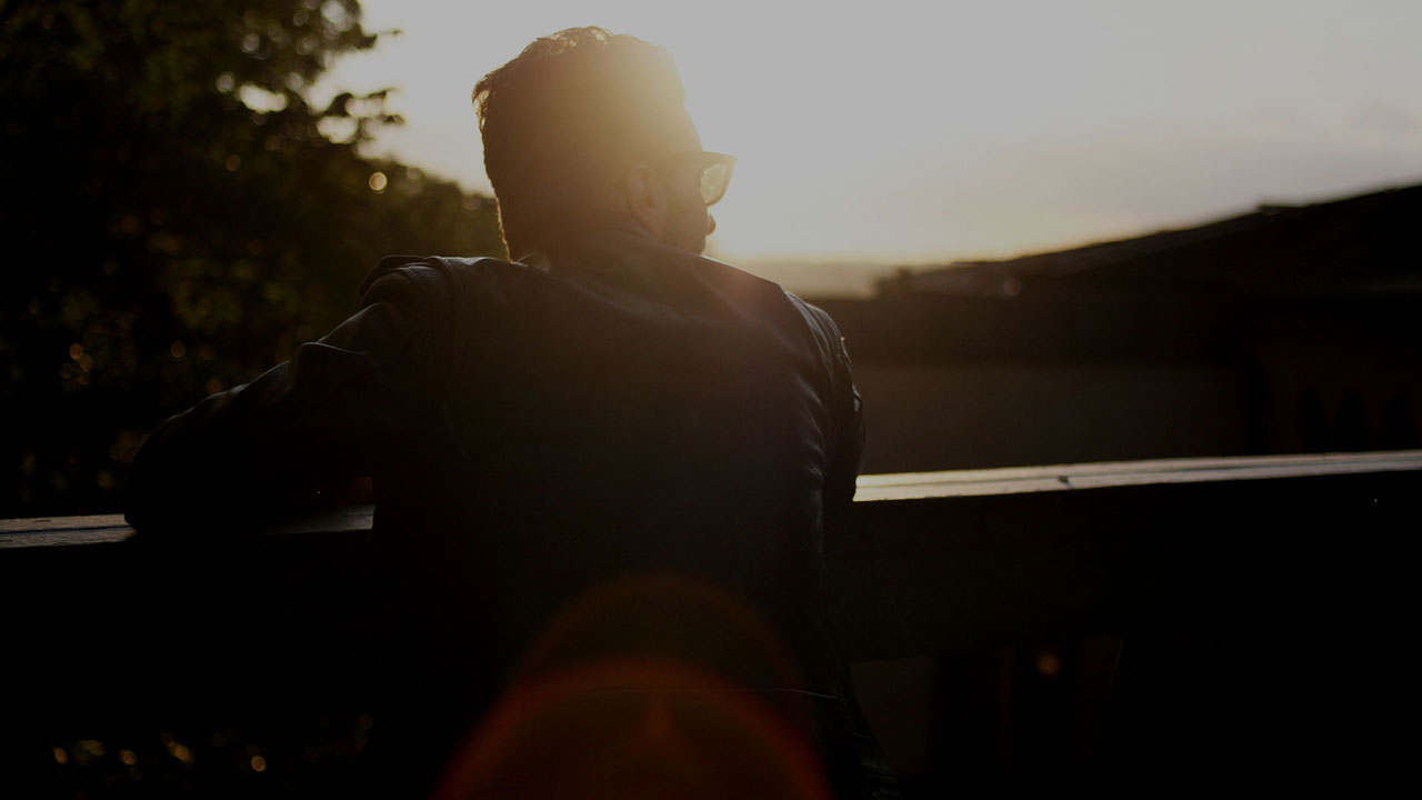 A man with sunglasses standing on a balcony infront of a bright sun