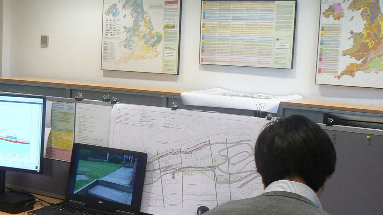 A man at a desk with two screens and a map, engaged in tasks that require both technology and geographical reference