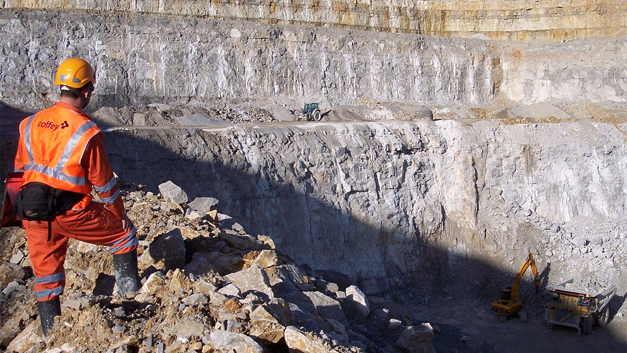 A man in an orange safety vest stands atop a cliff, surveying the construction site